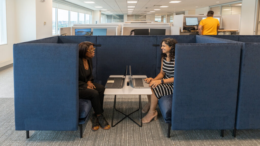 two women working on laptops and talking in padded cubicle