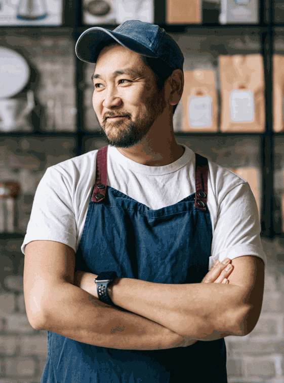 Asian man in hat and apron smiling while leaning in doorway src=