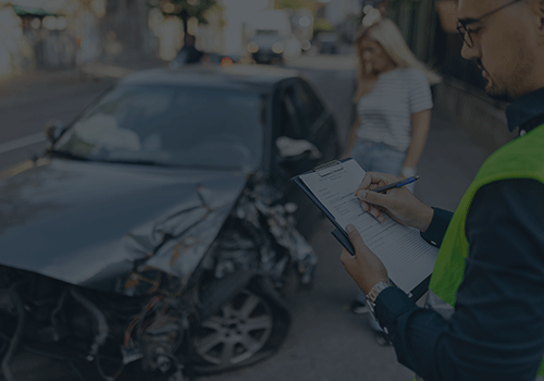 person logging details on clipboard with pen and paper with damaged car and driver standing in the background