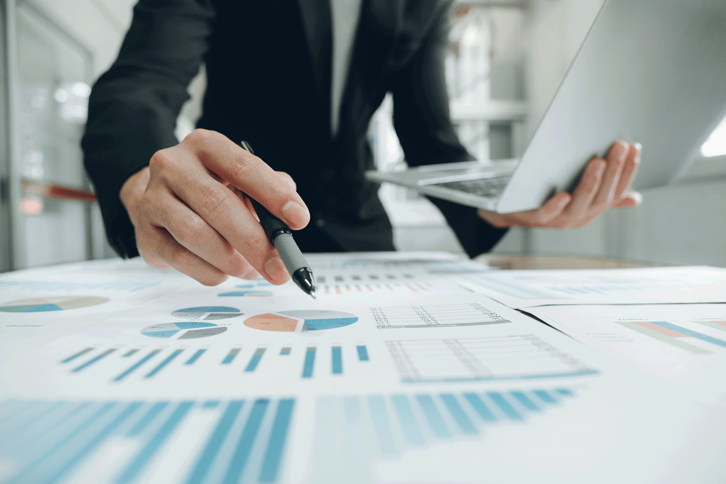 view from desk of paper graphs and person leaning over them holding a pen in their right hand and a laptop in their left hand