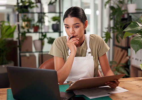 Female business owner reviewing laptop and clipboard and appearing deep in thought