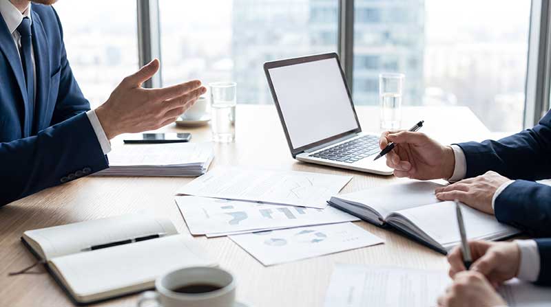focus on meeting desk with papers and laptop with people on edges.