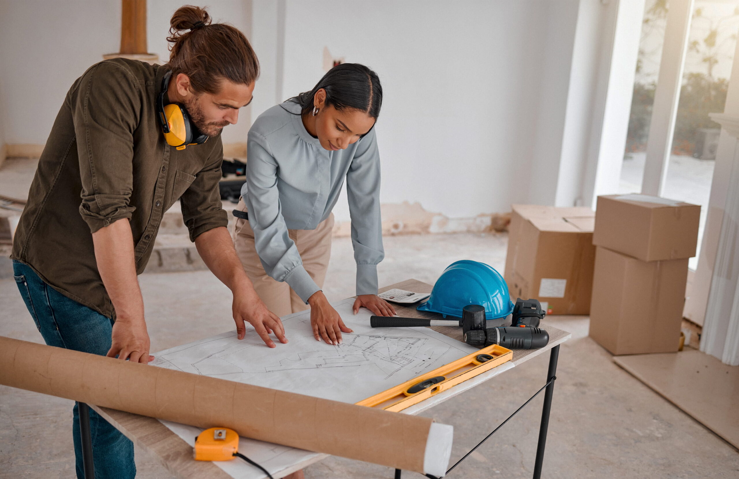 Couple planning a home renovation over blueprints, surrounded by tools, a hard hat, and moving boxes in a bright room.