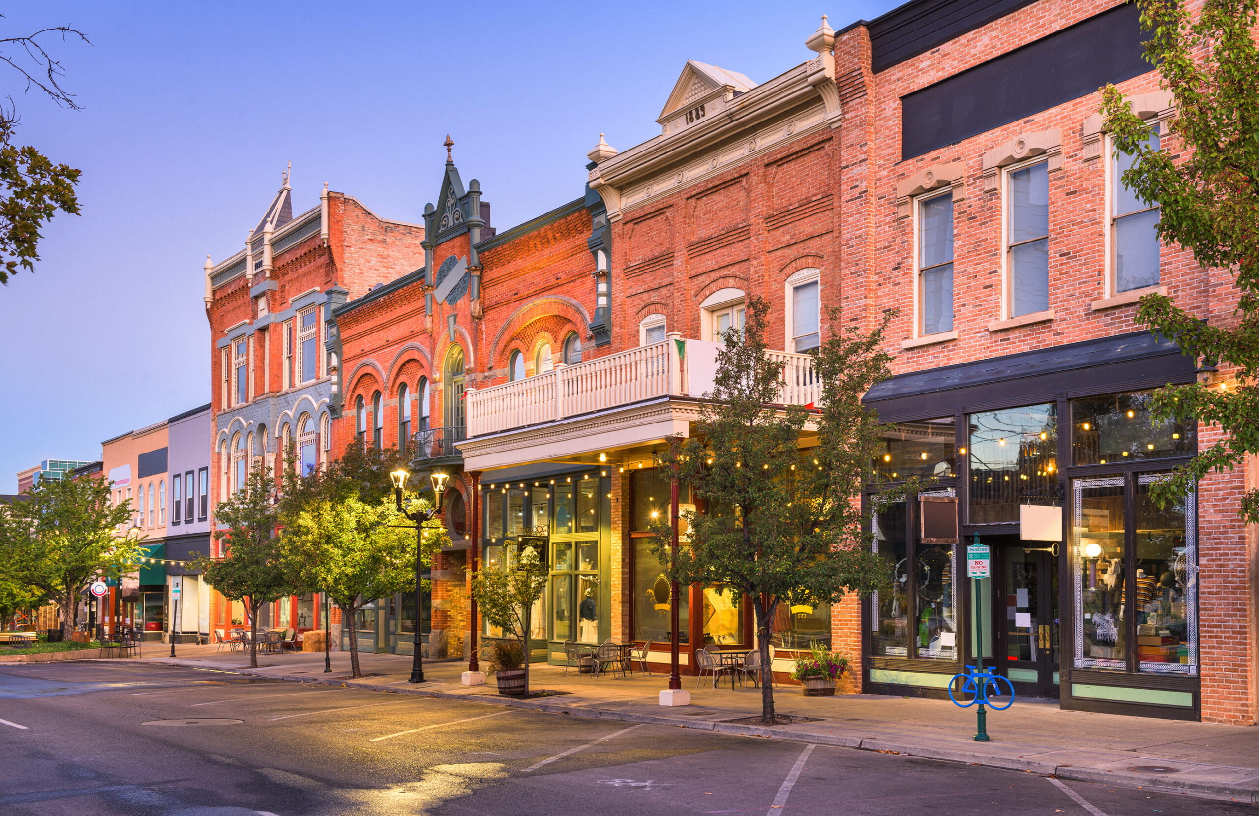 Historic red-brick downtown storefronts line a quiet street at dusk, with ornate architecture, glowing shop windows, trees, and warm lights.