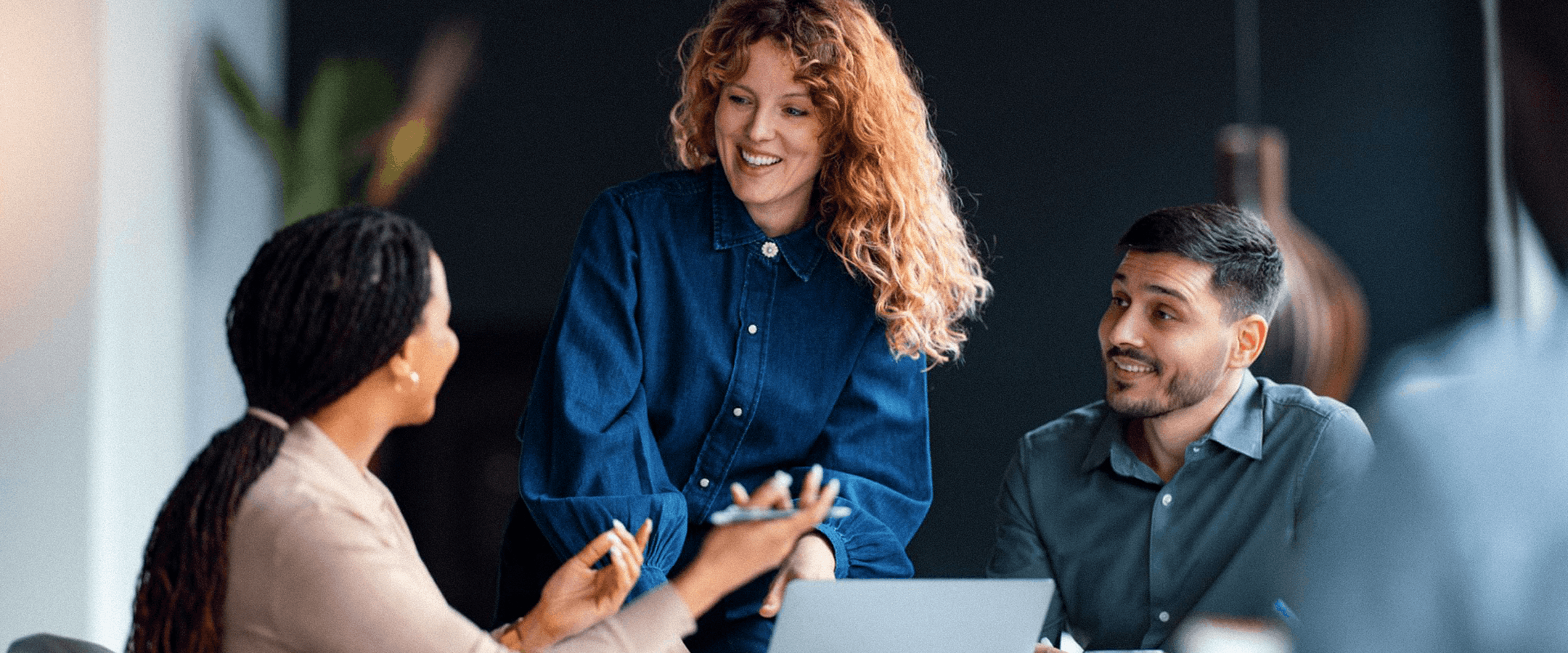 Three diverse coworkers collaborate in a modern office, smiling and discussing ideas around a laptop in a friendly, productive meeting.