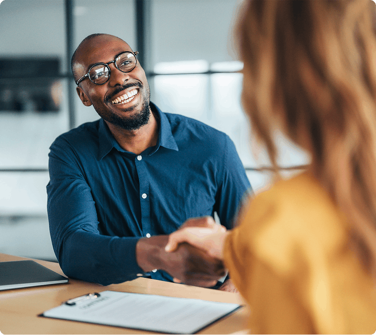 Smiling Black businessman in glasses shakes hands with a woman across a desk during a friendly, professional meeting in a bright office.