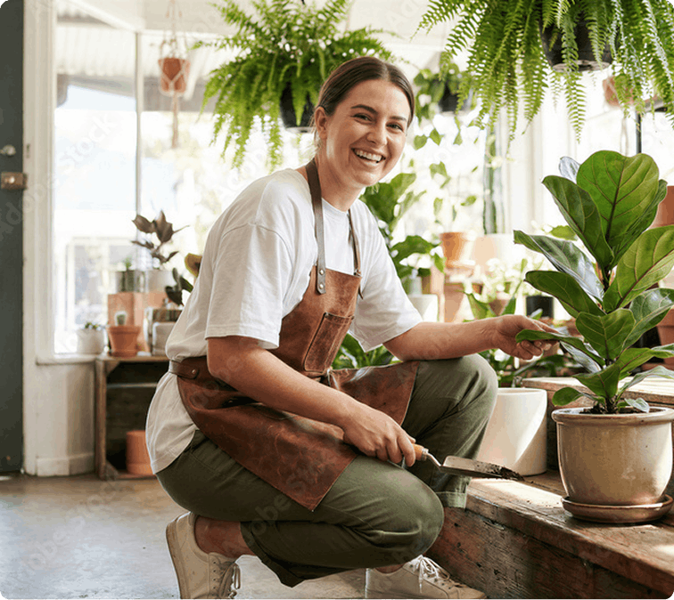 A woman in a brown apron kneels in a bright plant shop, smiling as she tends to a potted plant. Hanging ferns and natural light create a serene atmosphere.